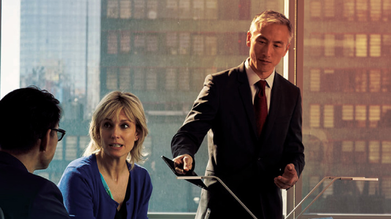 Three people are in an office meeting. A man in a suit is standing, pointing at a tablet, while a woman and another man sit at a table with coffee mugs and laptops. Sunlight filters through a large window behind them.