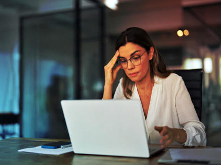 A woman in glasses sits at a desk in an office, concentrating on her laptop. She has a thoughtful expression, with one hand touching her forehead. Papers, a pen, and a smartphone are on the table. The background shows modern office decor.