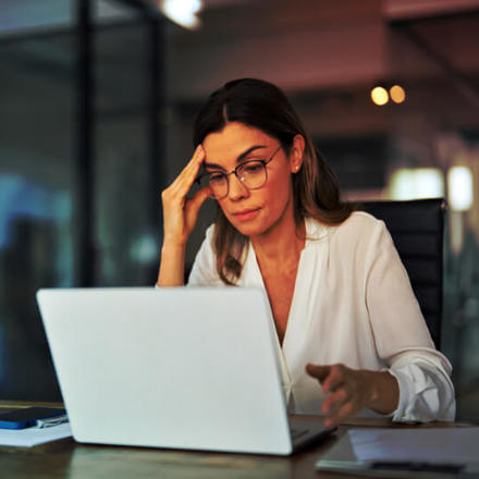 A woman in glasses sits at a desk in an office, concentrating on her laptop. She has a thoughtful expression, with one hand touching her forehead. Papers, a pen, and a smartphone are on the table. The background shows modern office decor.