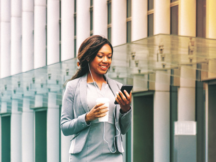 A woman in a business suit smiles while looking at her smartphone. She wears earphones and holds a takeaway coffee cup. The background features modern glass architecture, suggesting an urban environment.