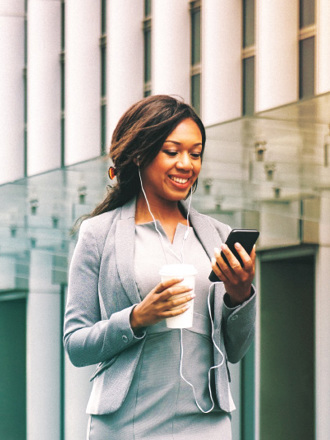 A woman in a business suit smiles while looking at her smartphone. She wears earphones and holds a takeaway coffee cup. The background features modern glass architecture, suggesting an urban environment.