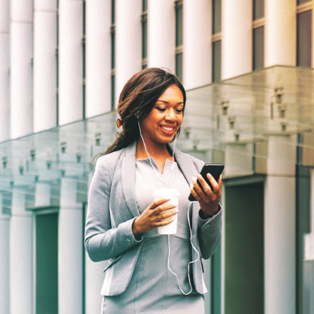 A woman in a business suit smiles while looking at her smartphone. She wears earphones and holds a takeaway coffee cup. The background features modern glass architecture, suggesting an urban environment.