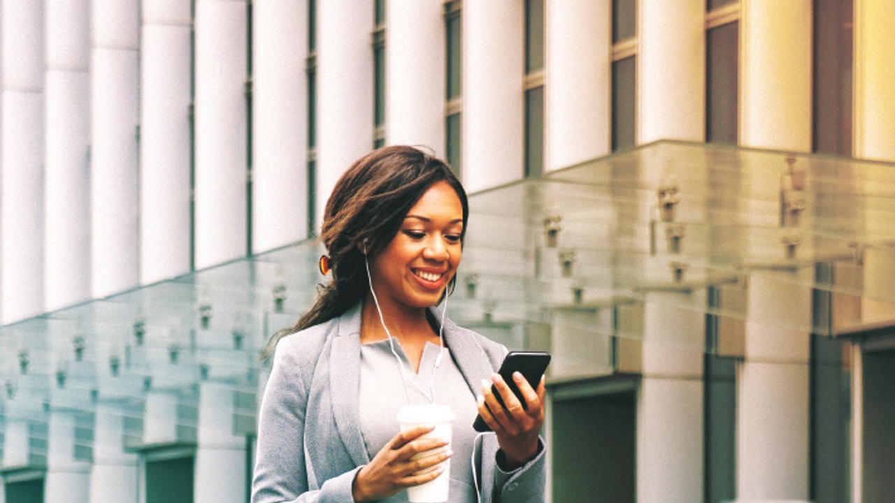 A woman in a business suit smiles while looking at her smartphone. She wears earphones and holds a takeaway coffee cup. The background features modern glass architecture, suggesting an urban environment.