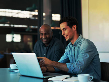 Two men are sitting at a table with a laptop, smiling and discussing something on the screen. One is pointing at the laptop. There are notebooks and coffee cups on the table. They appear to be in an office setting.