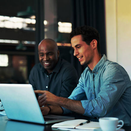 Two men are sitting at a table with a laptop, smiling and discussing something on the screen. One is pointing at the laptop. There are notebooks and coffee cups on the table. They appear to be in an office setting.