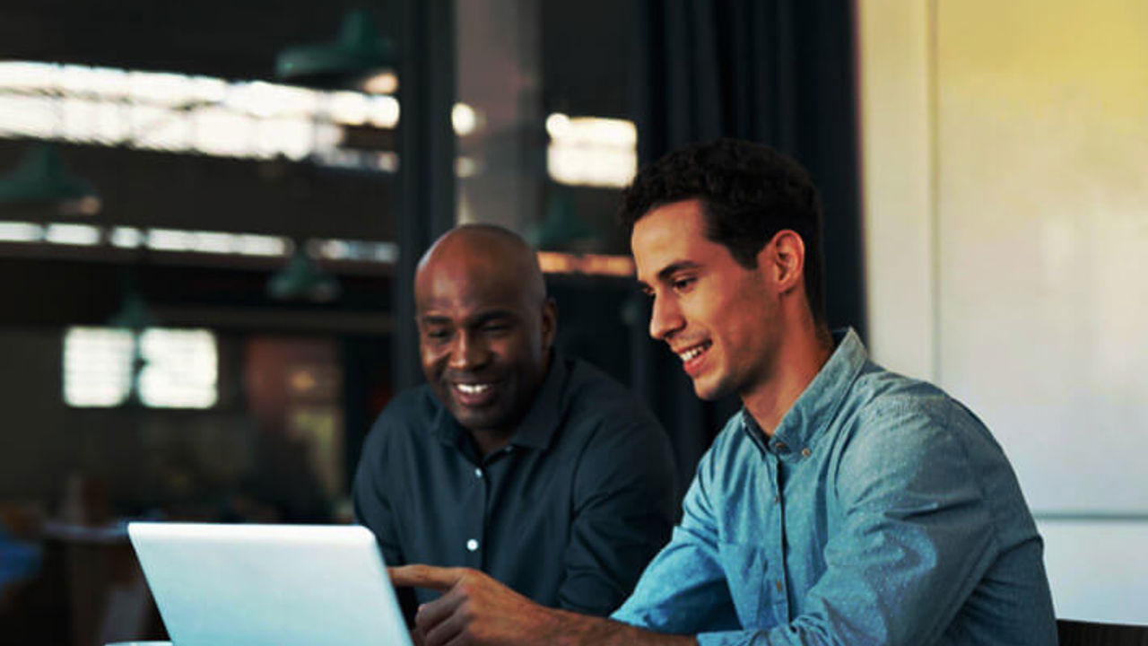 Two men are sitting at a table with a laptop, smiling and discussing something on the screen. One is pointing at the laptop. There are notebooks and coffee cups on the table. They appear to be in an office setting.