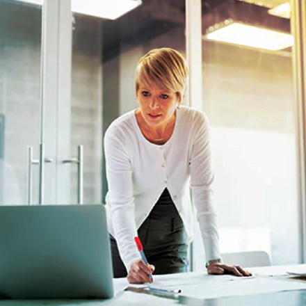 A person with short blonde hair stands at a desk, focused on a laptop and taking notes with a red pen. The setting is a modern office with glass doors in the background.