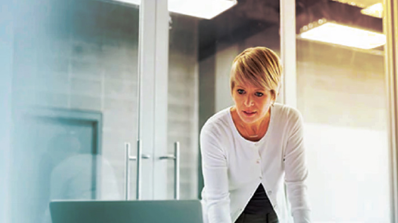 A person with short blonde hair stands at a desk, focused on a laptop and taking notes with a red pen. The setting is a modern office with glass doors in the background.