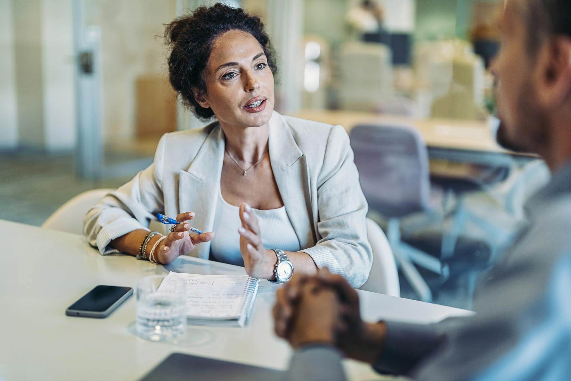 A woman in a blazer is having a discussion with a man across a table in an office setting. She gestures with her hands while speaking. A notepad, smartphone, and glass of water are on the table.